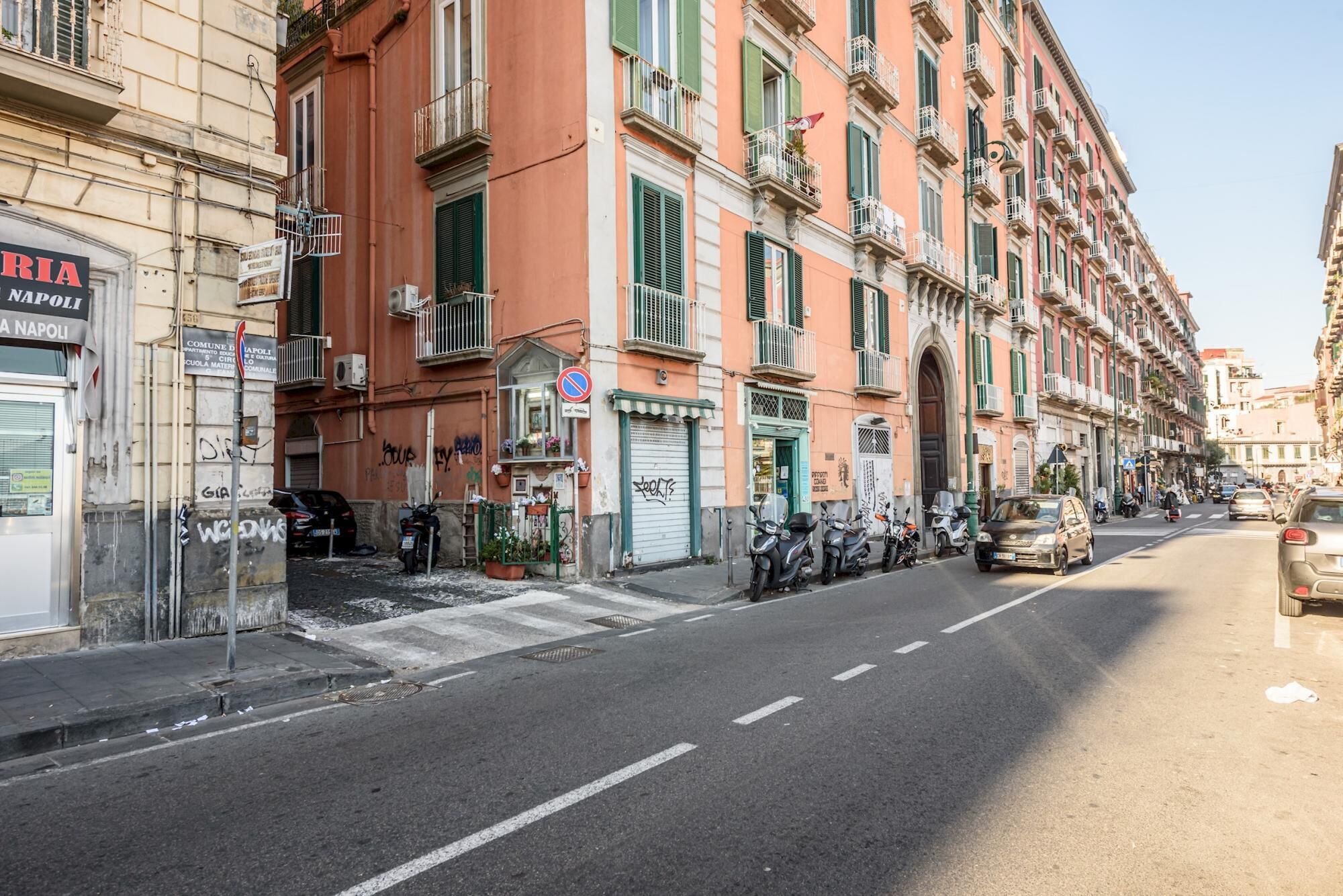 A street view with multi-story buildings, parked scooters, and a few cars.