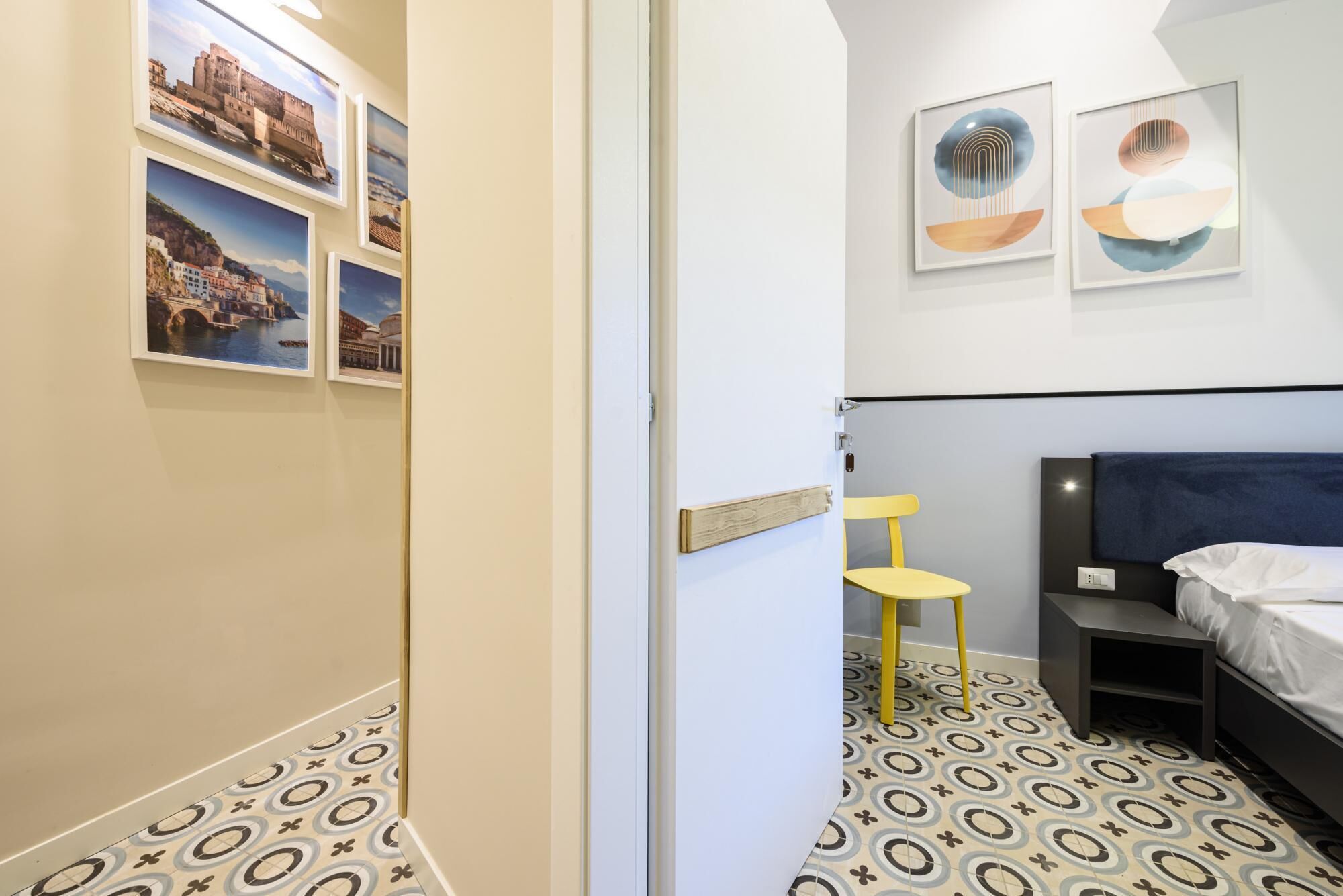 A hotel room with a bed, a chair, a small table, and a wall with framed pictures.