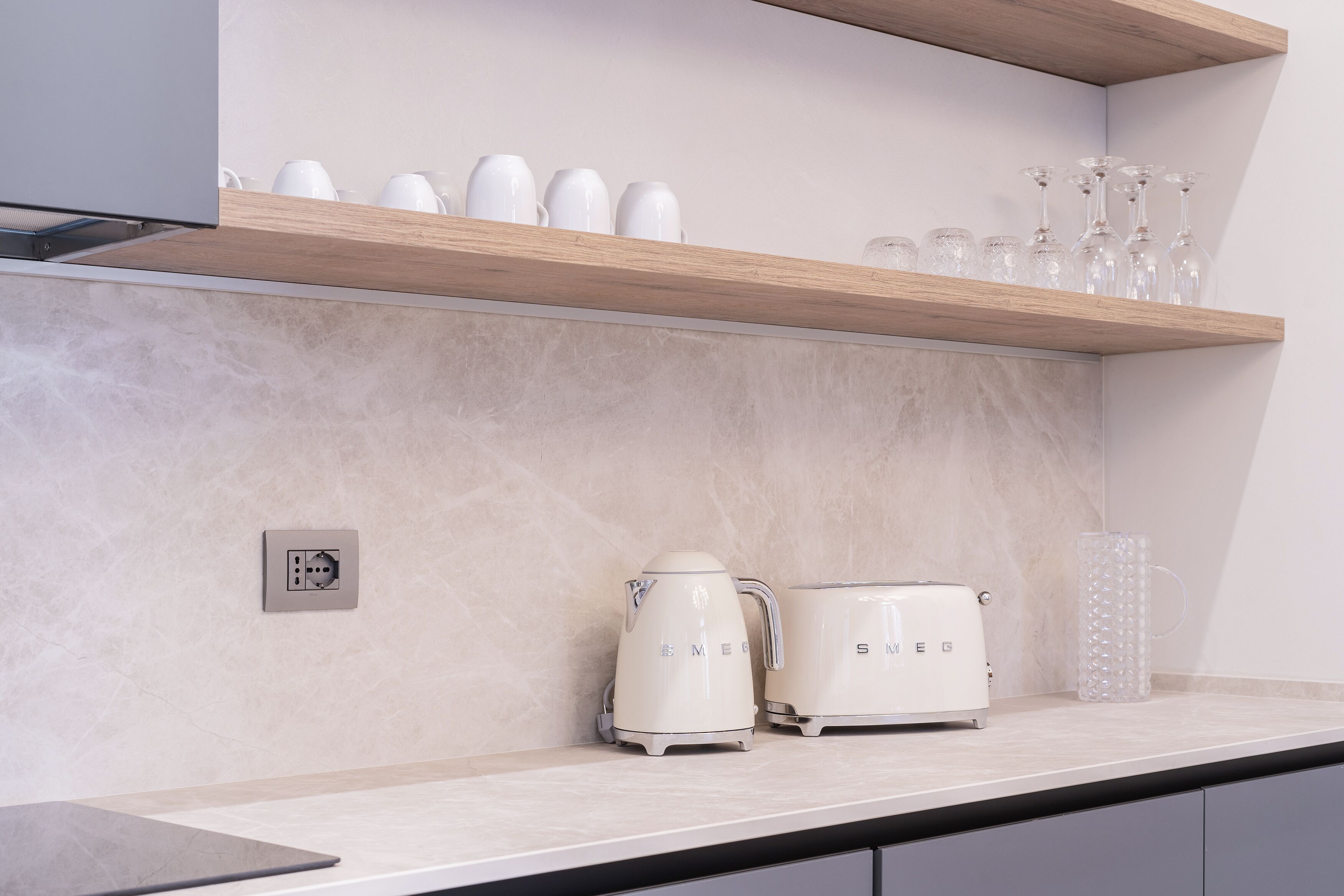 Una cocina con un estante de madera que contiene tazas y vasos blancos, una tetera eléctrica blanca y un tostador blanco.