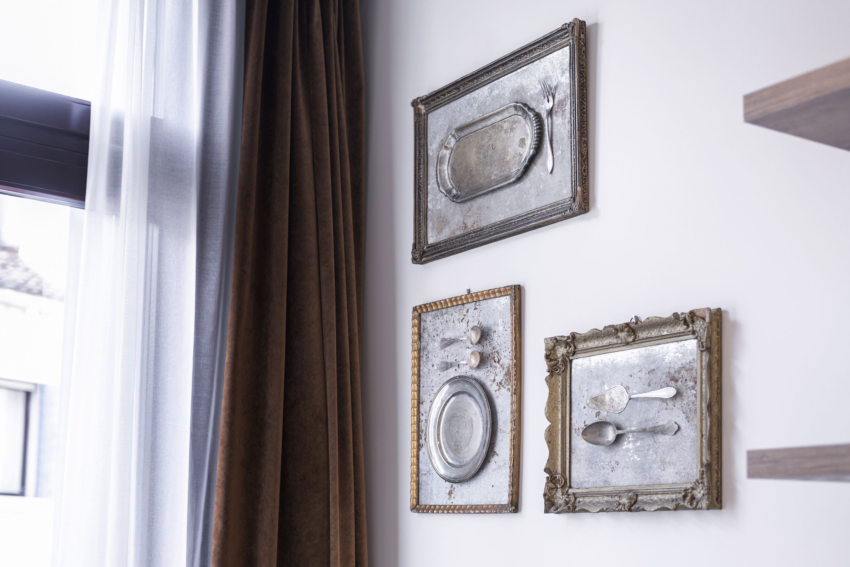 A wall with three vintage metal trays, each containing a spoon and a fork, and a window with curtains.
