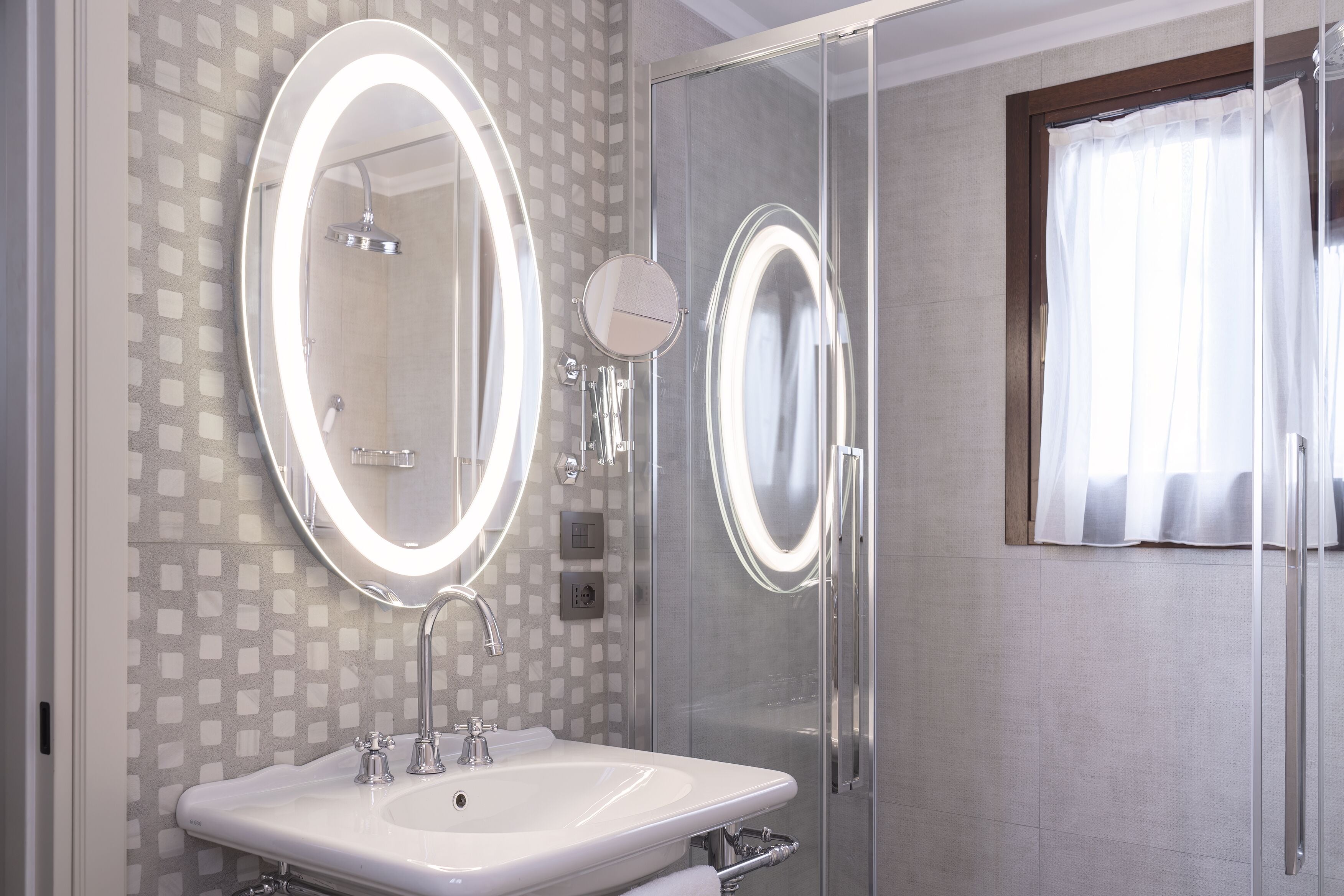 A bathroom with a large mirror, a sink, a shower, and a window with curtains.