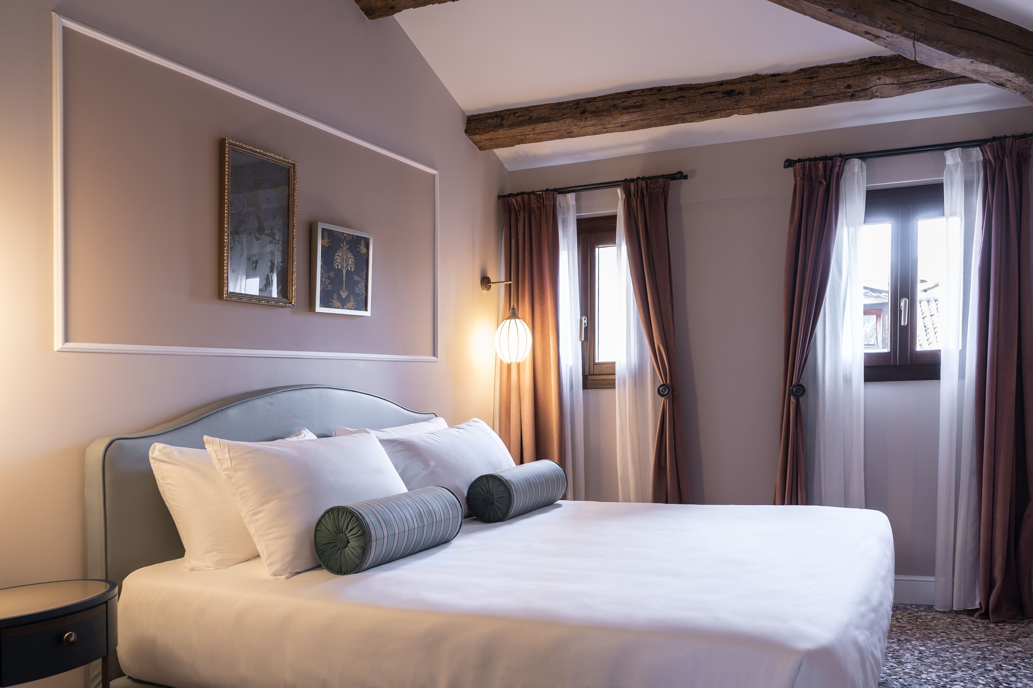 Una habitación de hotel con cama, mesita de noche, lámpara de pared y ventana con cortinas.