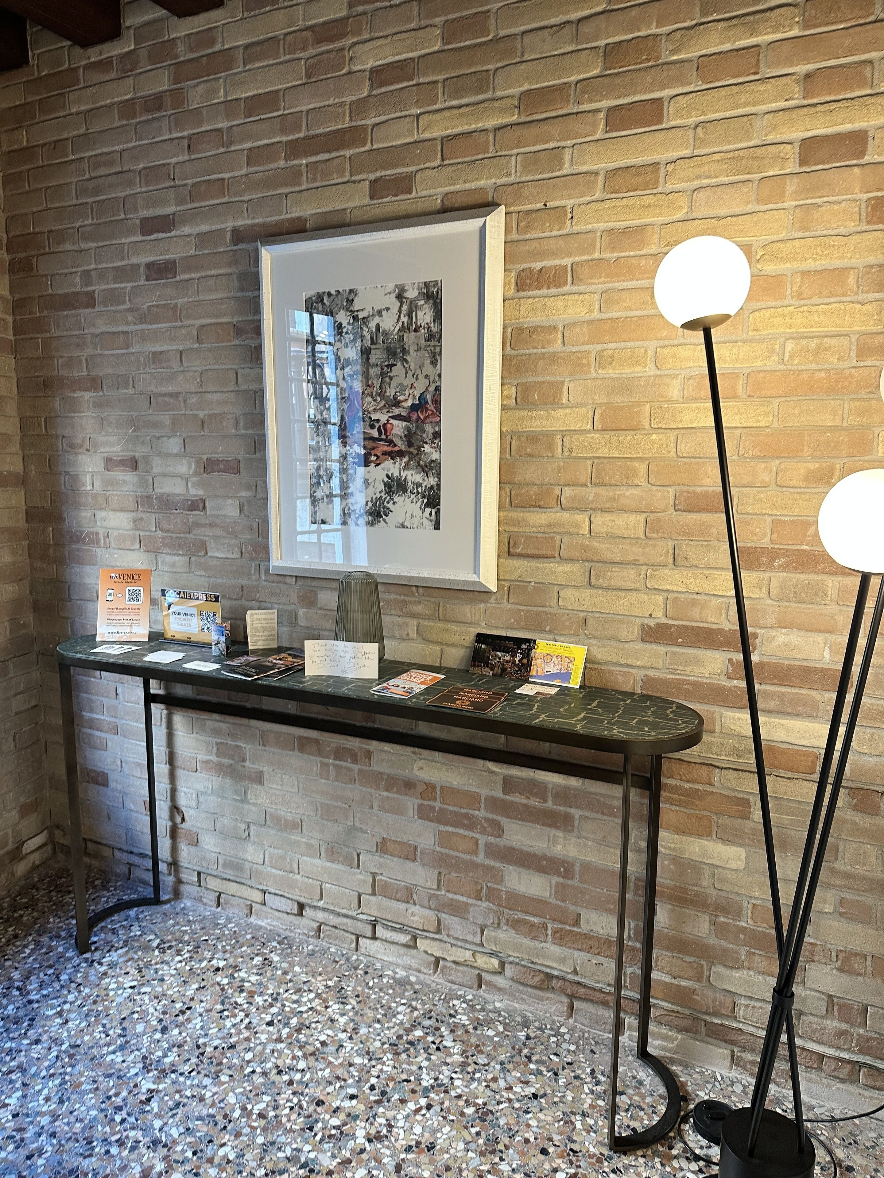 A reception area with a brick wall, a framed abstract painting, a reception desk with informational cards, and a tall floor lamp.