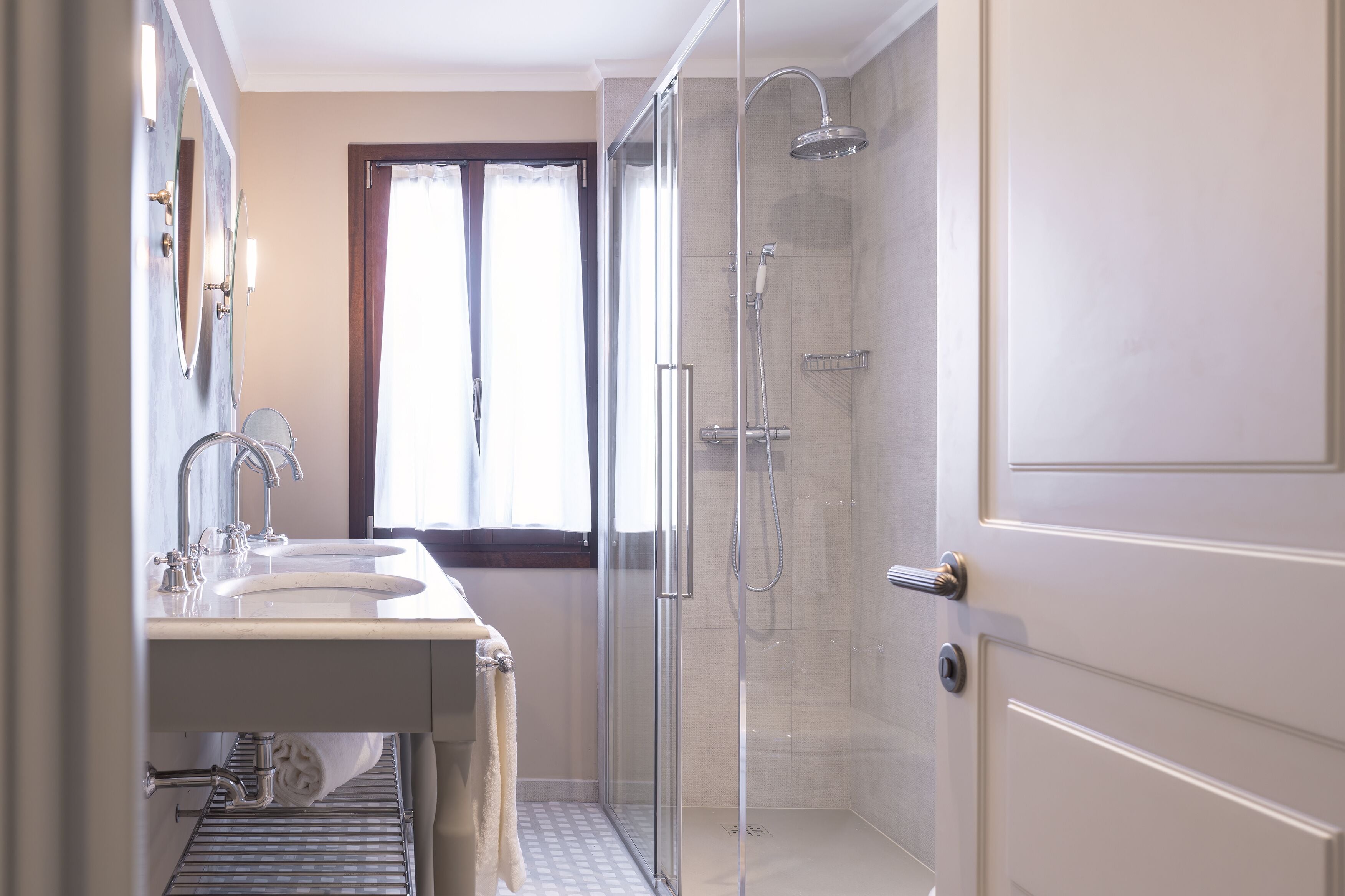 Une salle de bain moderne avec une douche à l’italienne en verre, un meuble-lavabo à deux vasques et une fenêtre avec des rideaux.