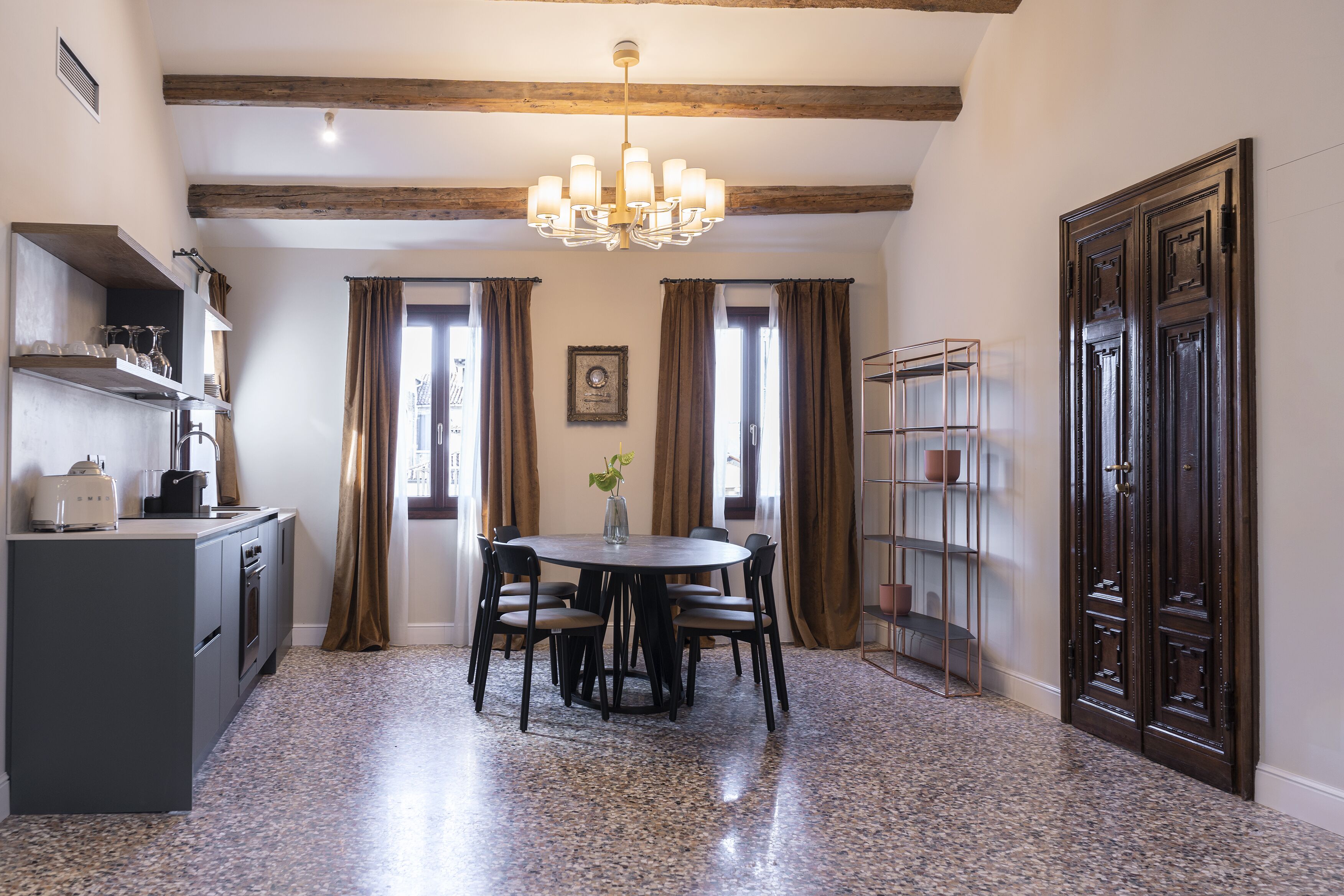 A dining area with a round table, chairs, a chandelier, and a kitchen area with appliances and shelves.