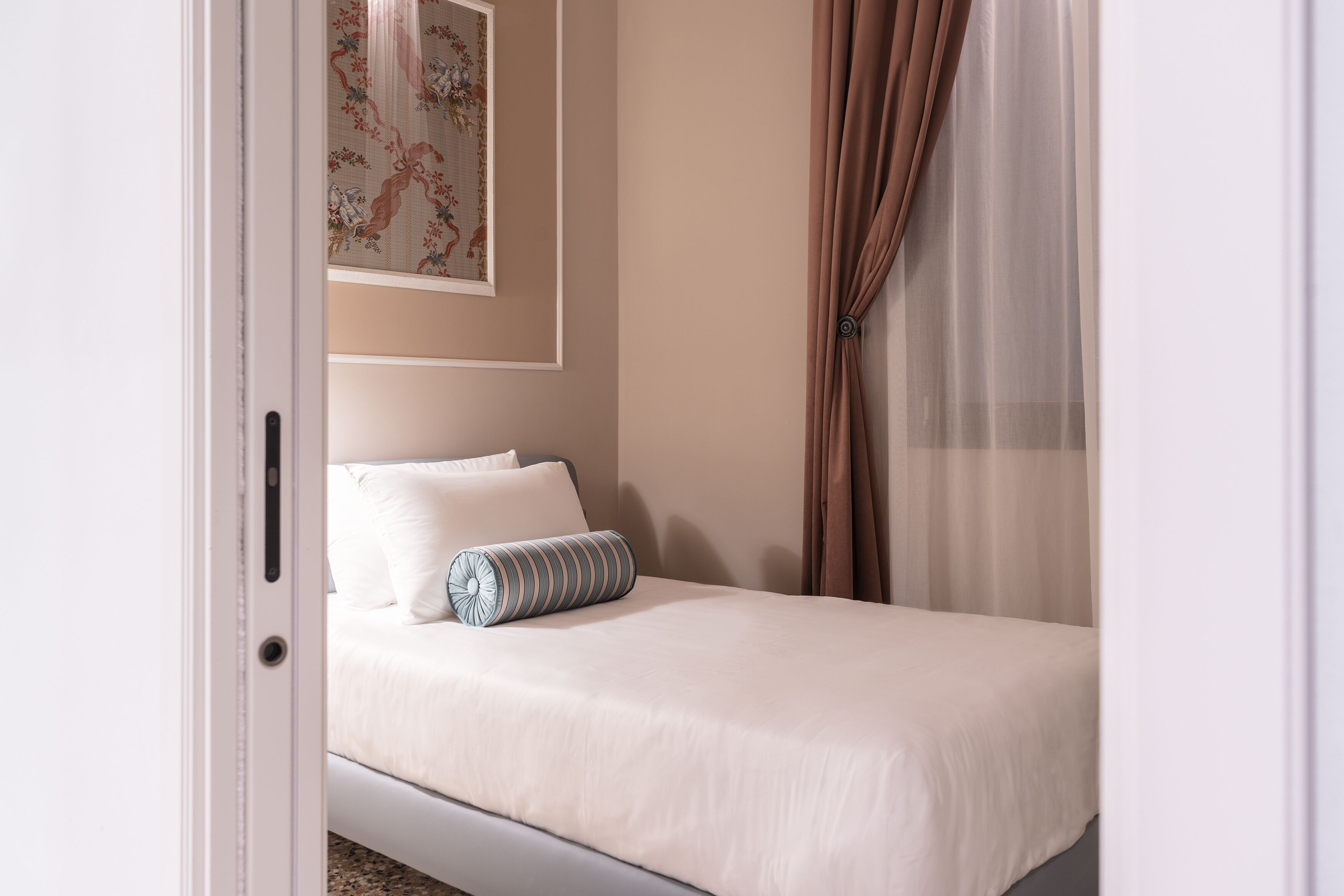 Una cama bien hecha con ropa blanca y una almohada a rayas, una obra de arte floral en la pared y una cortina entreabierta.