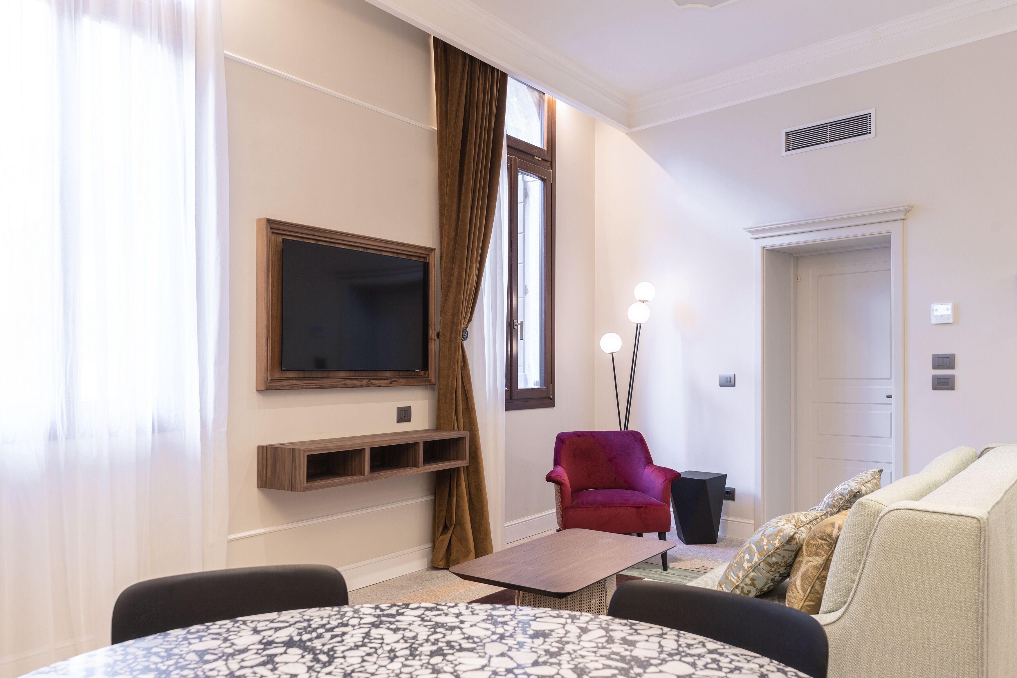 A hotel room with a flat-screen TV, a red armchair, a sofa, a small table, and a door.