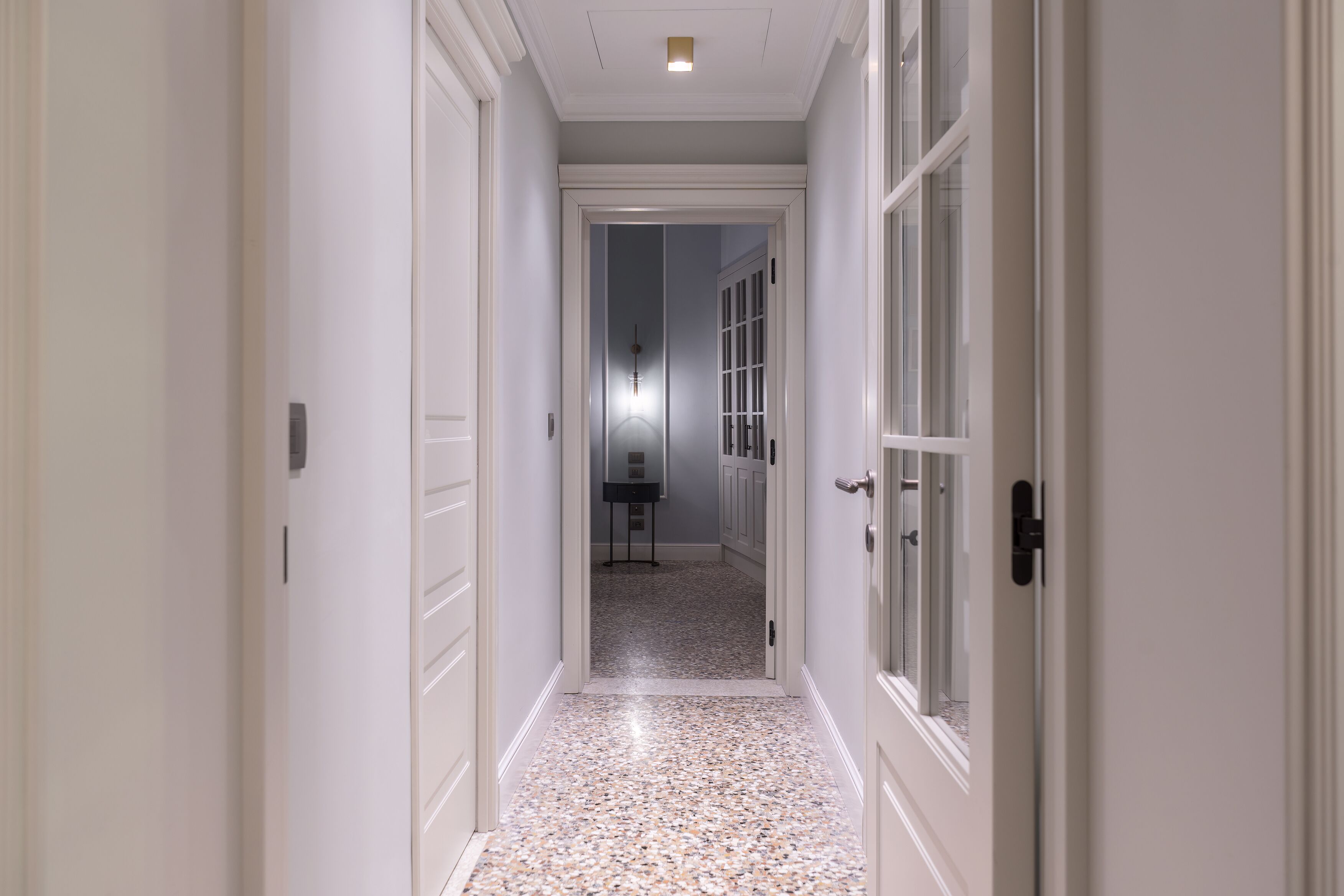Un pasillo con puertas blancas y un suelo moteado que conduce a una habitación con un escritorio y una silla.