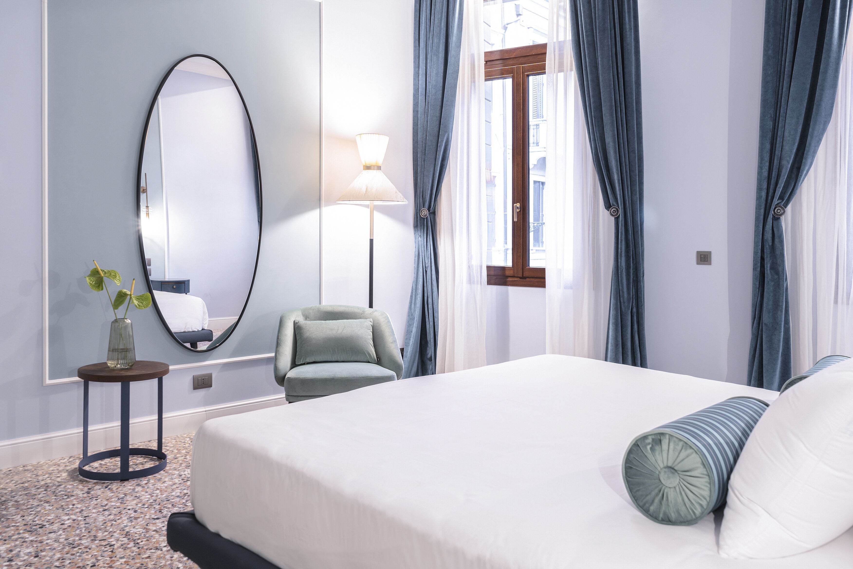 Une chambre d’hôtel avec un grand lit, un miroir rond, une petite table avec une plante, une chaise et une fenêtre avec des rideaux.