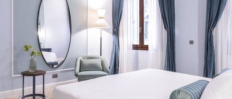 Une chambre d’hôtel avec un grand lit, un miroir rond, une petite table avec une plante, une chaise et une fenêtre avec des rideaux.