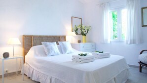 A neatly arranged bedroom with a large bed, a bedside table with a lamp, a small bedside table with a decorative item, a nightstand, and a window with curtains. - The Saints - St Mathias 2 Bedroom Cottage (Corfu)