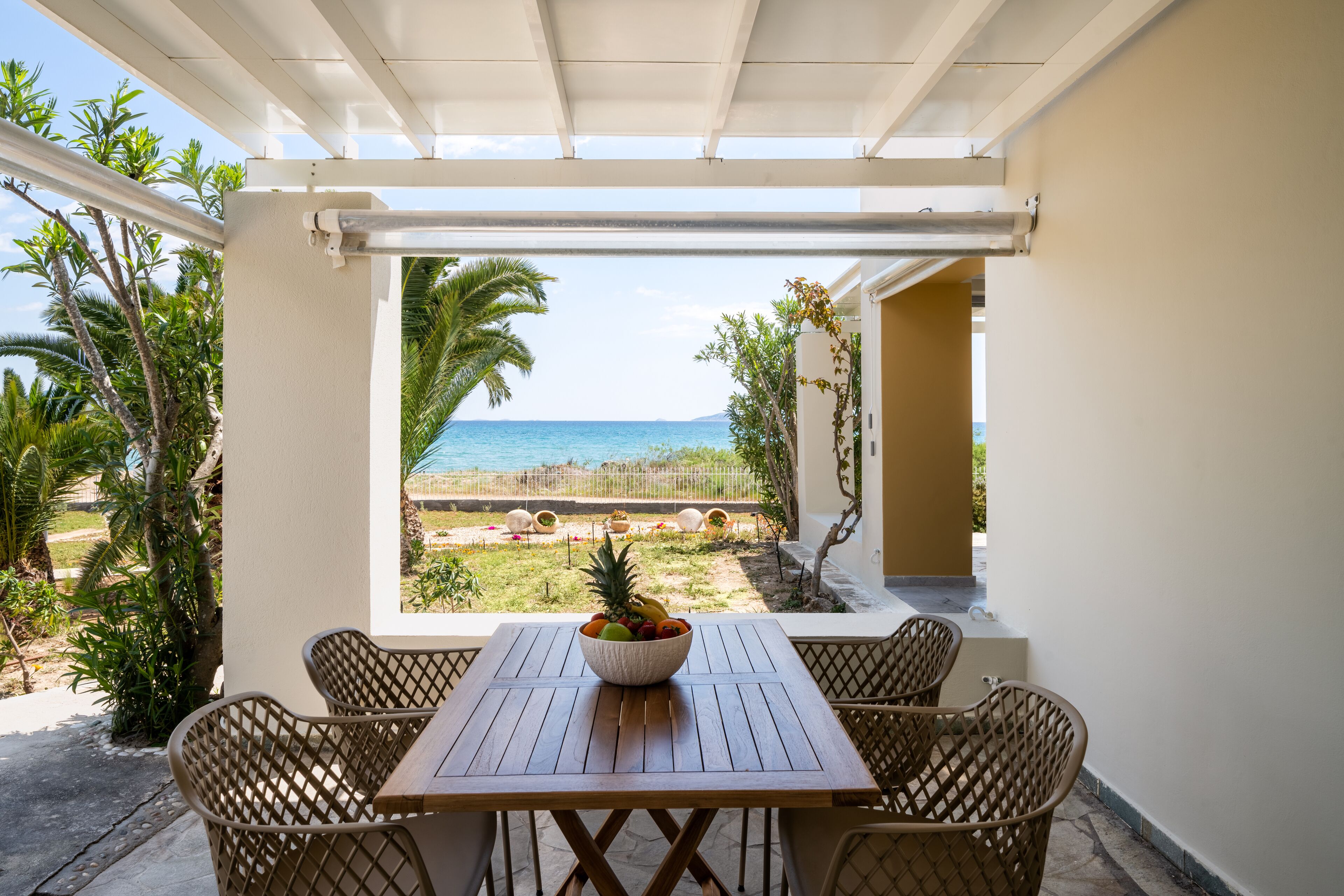 A covered patio with a wooden table and wicker chairs, overlooking a beach and ocean.
