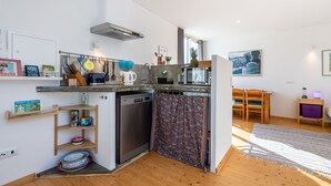 A compact kitchen with a dishwasher, microwave, and a small refrigerator. There is a wooden chair and a small table with a lamp.
