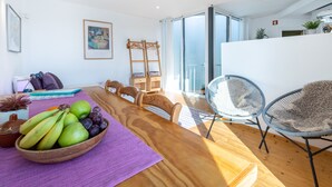 A modern living room with a wooden dining table, purple table runner, and a bowl of fruit. There are two chairs, a sofa, and a large window with a view of the outdoors.