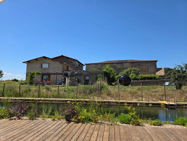 A rustic stone building with a wooden deck and a pond. - Nature gite, natural swimming pool, sauna, Nordic bath, farmhouse (Augerolles)