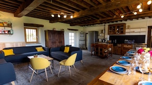 Uma sala de estar aconchegante, com um sofá azul escuro e almofadas amarelas, uma mesa de jantar de madeira para seis pessoas e uma área de cozinha com armários de madeira e uma geladeira.