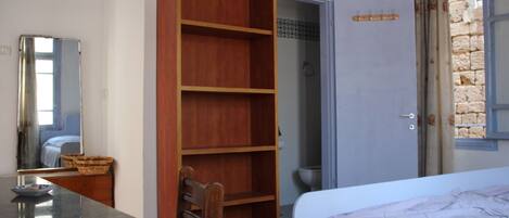A bedroom with a bed, a wooden wardrobe, a bathroom with a shower, and a window with a view of stone buildings.
