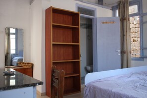 A bedroom with a bed, a wooden wardrobe, a bathroom with a shower, and a window with a view of stone buildings. - Nzar Khoury For Hosting (Acre)