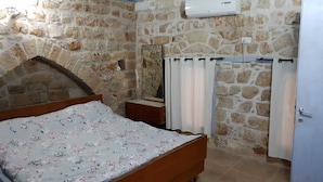 A bedroom with a stone wall, a wooden bed, a nightstand, a mirror, a white air conditioning unit, and a door. - Nzar Khoury For Hosting (Acre)