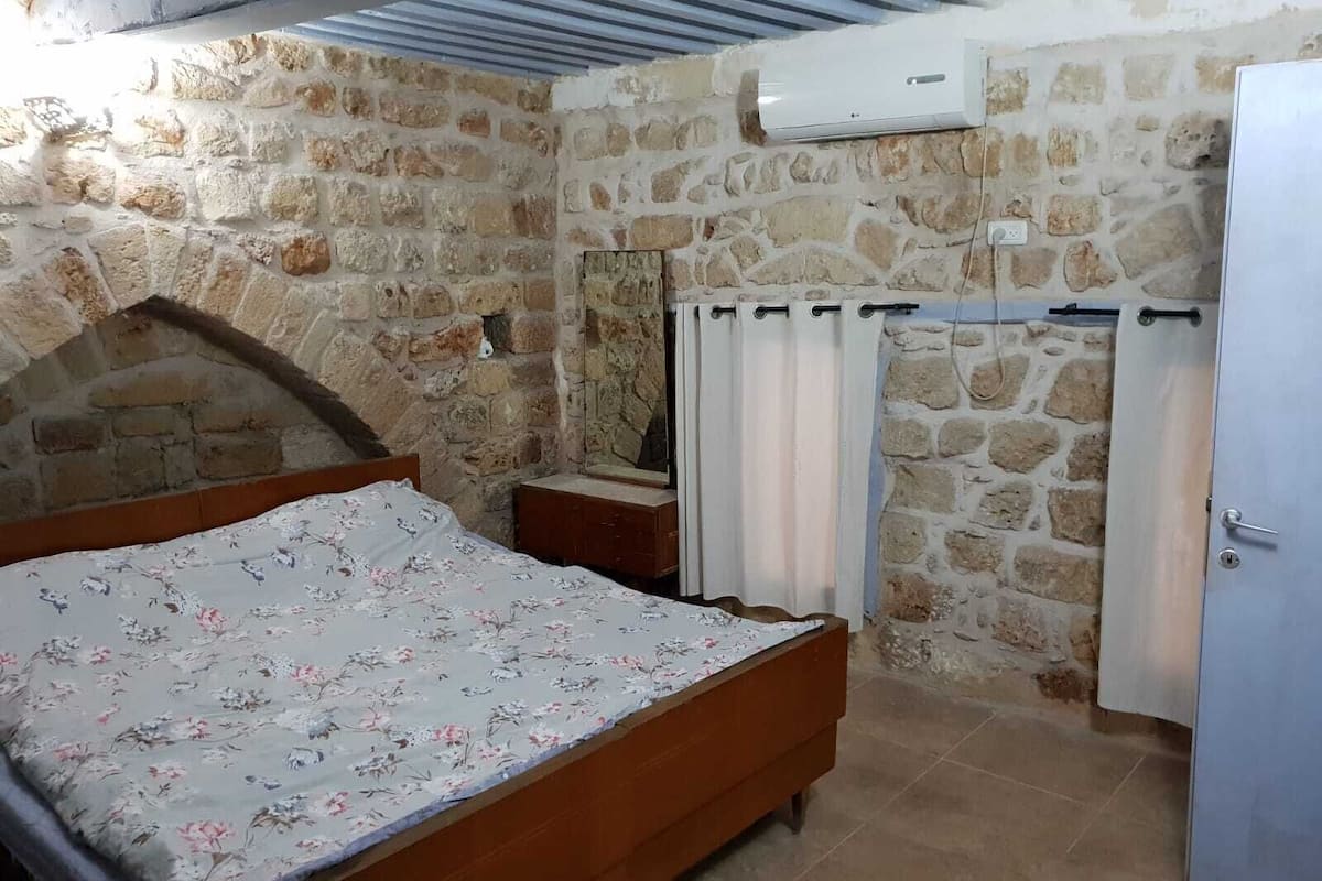 Un dormitorio con pared de piedra, una cama de madera, una mesita de noche, un espejo, un equipo de aire acondicionado blanco y una puerta.