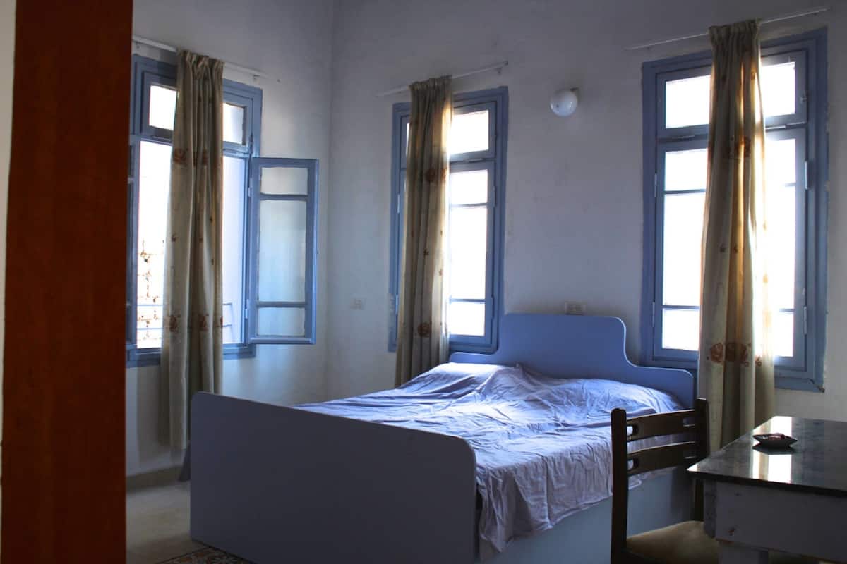 Habitación individual con una cama y cabecera de madera, una mesita y una ventana con cortinas.