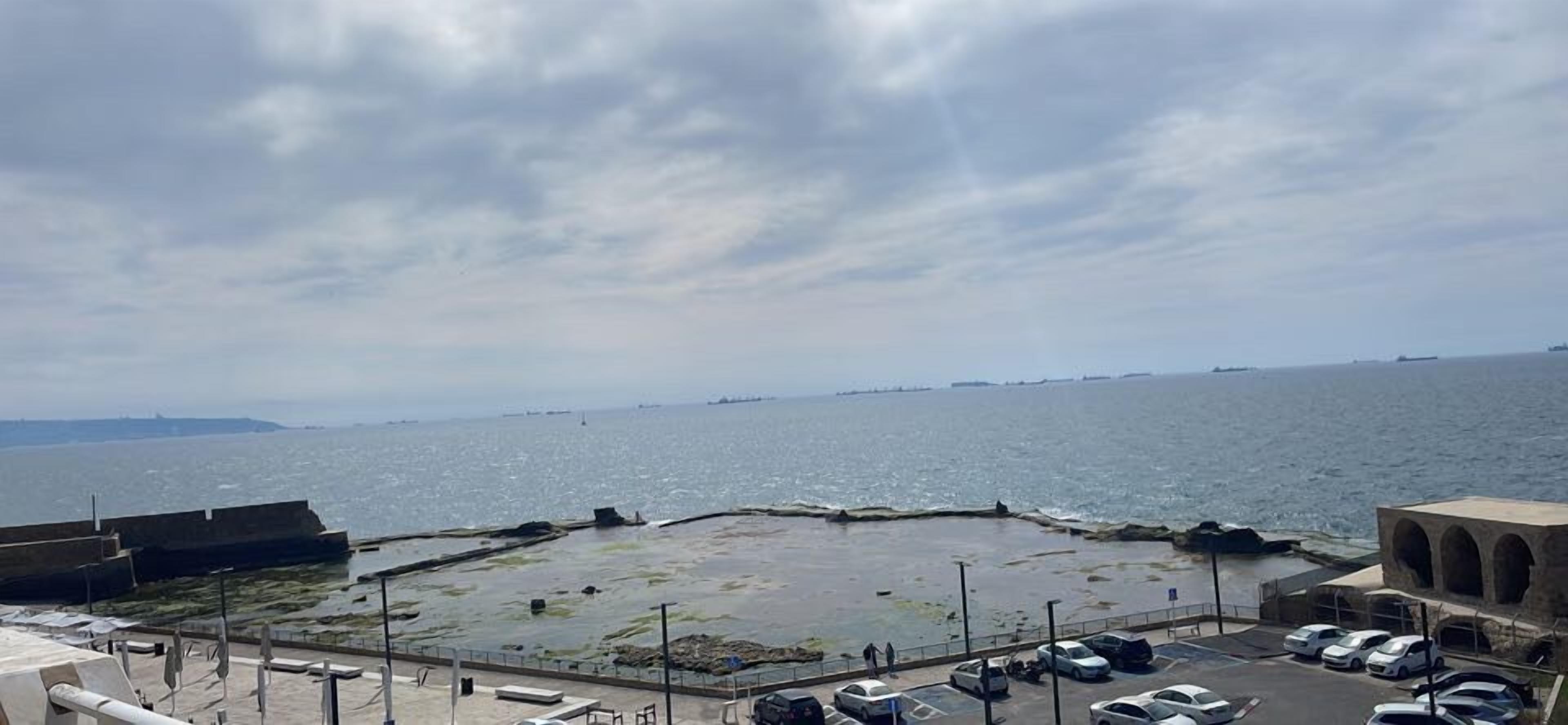 Vista costera con un rompeolas rocoso, un estacionamiento y un edificio con ventanas de arco.