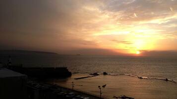 A sunset over a calm sea with a pier and parked cars.