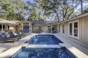 A pool area with lounge chairs, a covered patio, and a house with large windows.