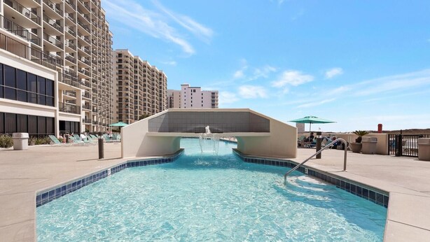 Une piscine agrémentée d’un élément décoratif aquatique au centre, entourée d’un espace aménagé avec des chaises longues et un bâtiment visible au loin.