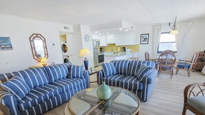 A living room with striped sofas, a glass coffee table, wicker chairs, a kitchen in the background, and a laundry area.
