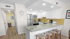 A modern kitchen with white cabinets, a stainless steel refrigerator, a built-in oven, and a granite countertop. There are three wooden bar stools and a clock on the wall.