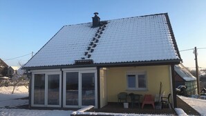 Pieni talo, jonka katto on lumen peitossa, ja lasiovet johtavat terassialueelle, jossa on ulkokalusteita.