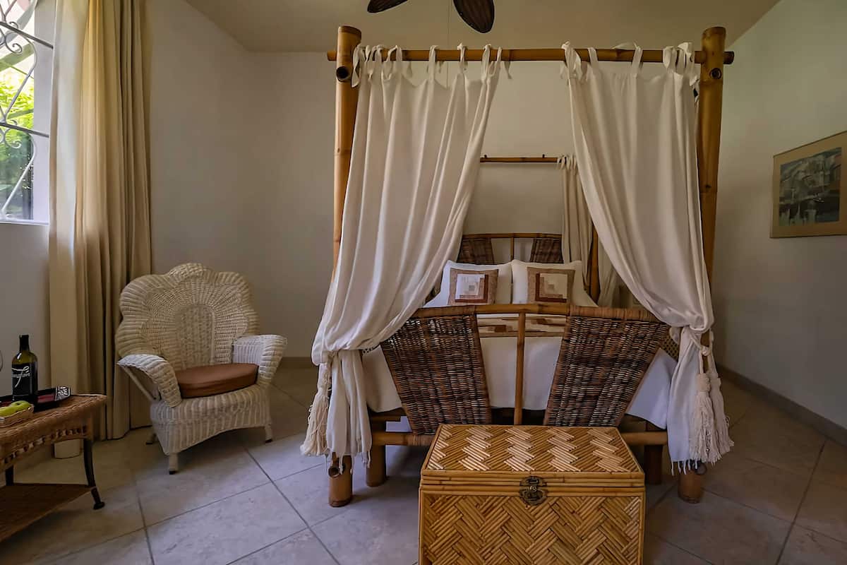 A room with a four-poster bed, wicker furniture, a small table with a bottle, and a framed picture on the wall.