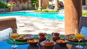 Zona de comedor junto a la piscina con macetas de terracota, platos de comida y vasos de zumo de naranja.