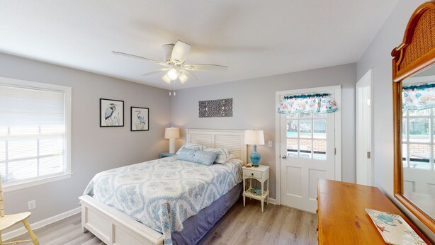 A bedroom with a bed, two bedside lamps, a ceiling fan, a window with blinds, a door with a decorative curtain, and a small wooden table. - Turtle's Nest Duplex 3 bedrooms, 2 bathrooms (Jekyll Island)