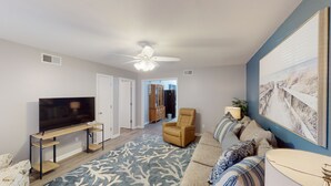 A living room with a TV, a ceiling fan, a sofa, and a painting on the wall. - Turtle's Nest Duplex 3 bedrooms, 2 bathrooms (Jekyll Island)
