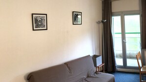 Une pièce avec un canapé marron, deux tableaux encadrés au mur, un lampadaire et une vue sur l’extérieur, visible à travers une porte vitrée.