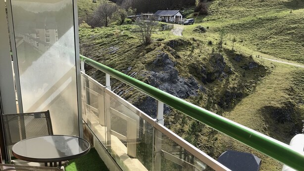 Un balcon doté d’une balustrade en verre, qui offre une vue sur une colline verdoyante, avec une maison et une voiture garée.