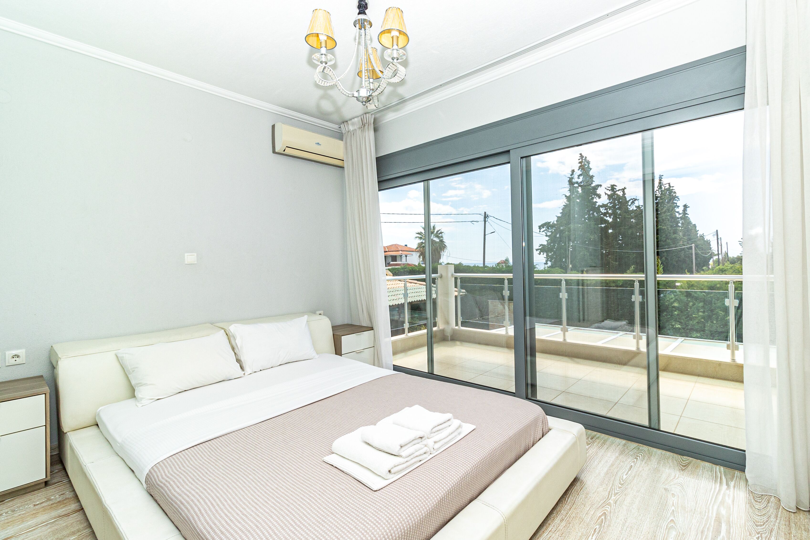 Un dormitorio moderno con una cama grande, una araña y un balcón con vistas a árboles y edificios.