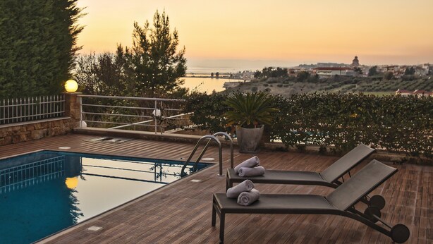 A wooden deck with a swimming pool, lounge chairs, and a view of the sea at sunset. - Villa Nefeli (Nea Propontida)