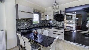 A modern kitchen with a dining area, featuring a black countertop, white chairs, and stainless steel appliances. - Villa Nefeli (Nea Propontida)