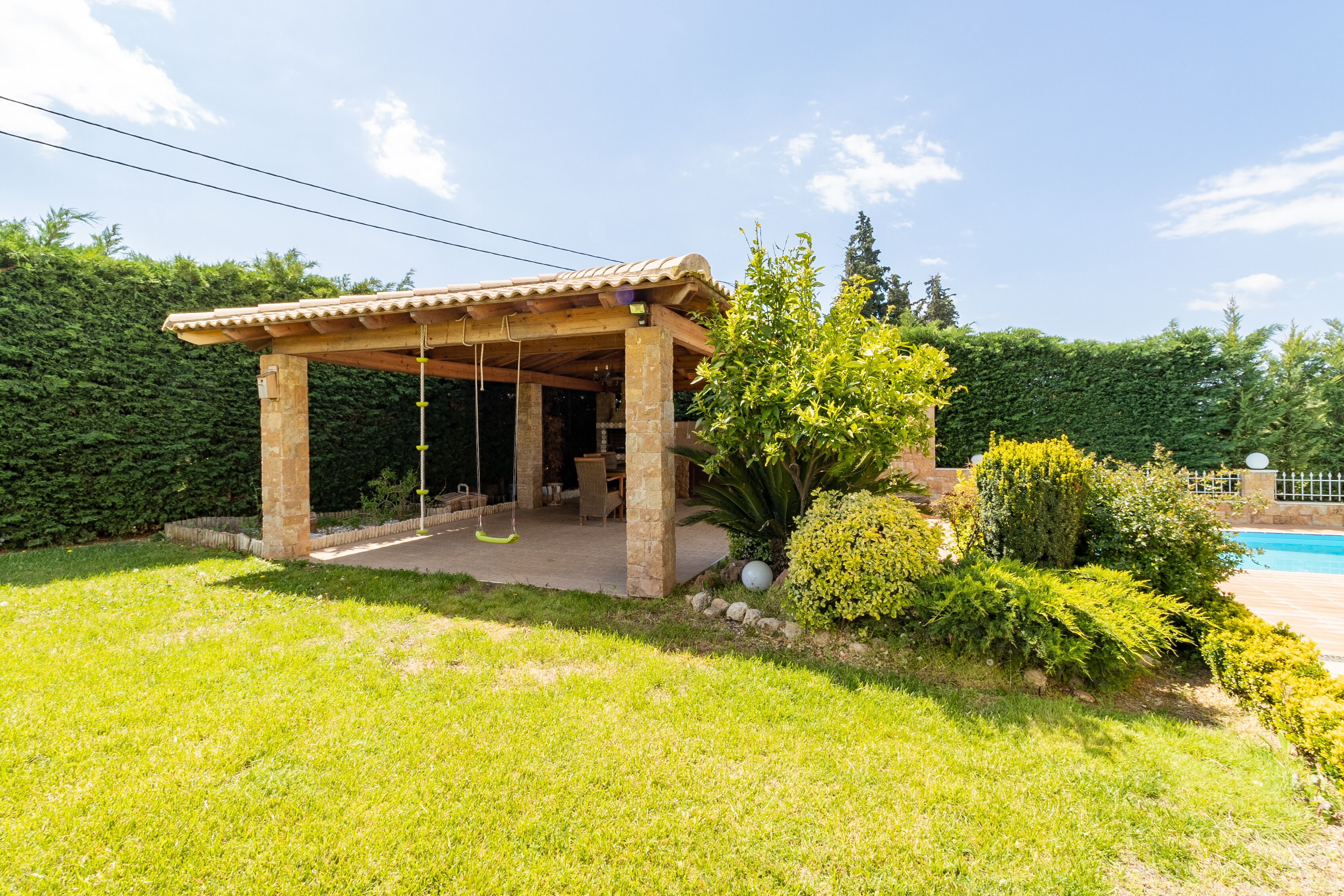 A covered outdoor seating area with a swing, surrounded by lush greenery and a pool.
