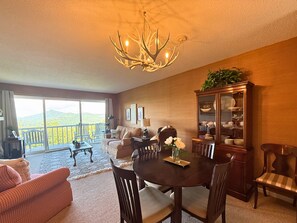 A dining area with a chandelier, a dining table, chairs, a cabinet with dishes, and a view of a balcony with a railing. - Cozy Mountain Condo with Mountain Views (Sugar Mountain)