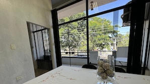 A dining area with a table covered in a floral tablecloth, a vase with decorative items, and a view of trees through large windows. - Loft São João Exclusive (Sao Joao da Boa Vista)
