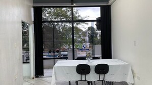 A dining area with a table set for two, large windows with curtains, and a view of the street outside. - Loft São João Exclusive (Sao Joao da Boa Vista)