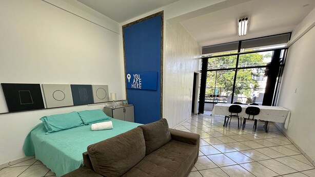 A hotel room with a bed, a brown sofa, a dining table with chairs, and a large window. - Loft São João Exclusive (Sao Joao da Boa Vista)