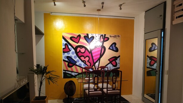 Une pièce avec un mur jaune, un grand tableau abstrait, un ventilateur et une chaise.