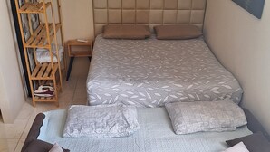 Un lit avec une couvre-lit à motifs, plusieurs oreillers et une étagère en bois sur laquelle se trouvent un vêtement plié et un fer à repasser.