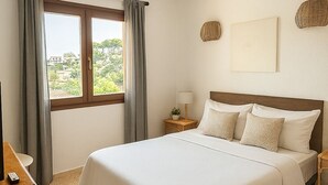 Une chambre d’hôtel avec un grand lit, une table de chevet avec une lampe, une petite table avec une plante et une fenêtre avec des rideaux.