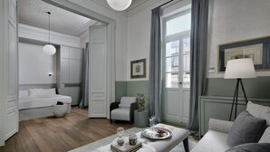 Ein modernes, elegant eingerichtetes Wohnzimmer mit Sofa, Sessel und Hocker, einem Esstisch mit Tablett sowie einem großen Fenster mit Vorhängen.